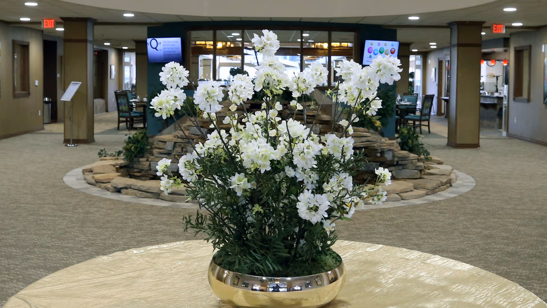 A large indoor common area with a round table in the foreground holding a gold vase filled with white flowers. In the background, there is a stone water feature surrounded by chairs and tables, with carpeted floors and multiple doorways leading to other rooms.