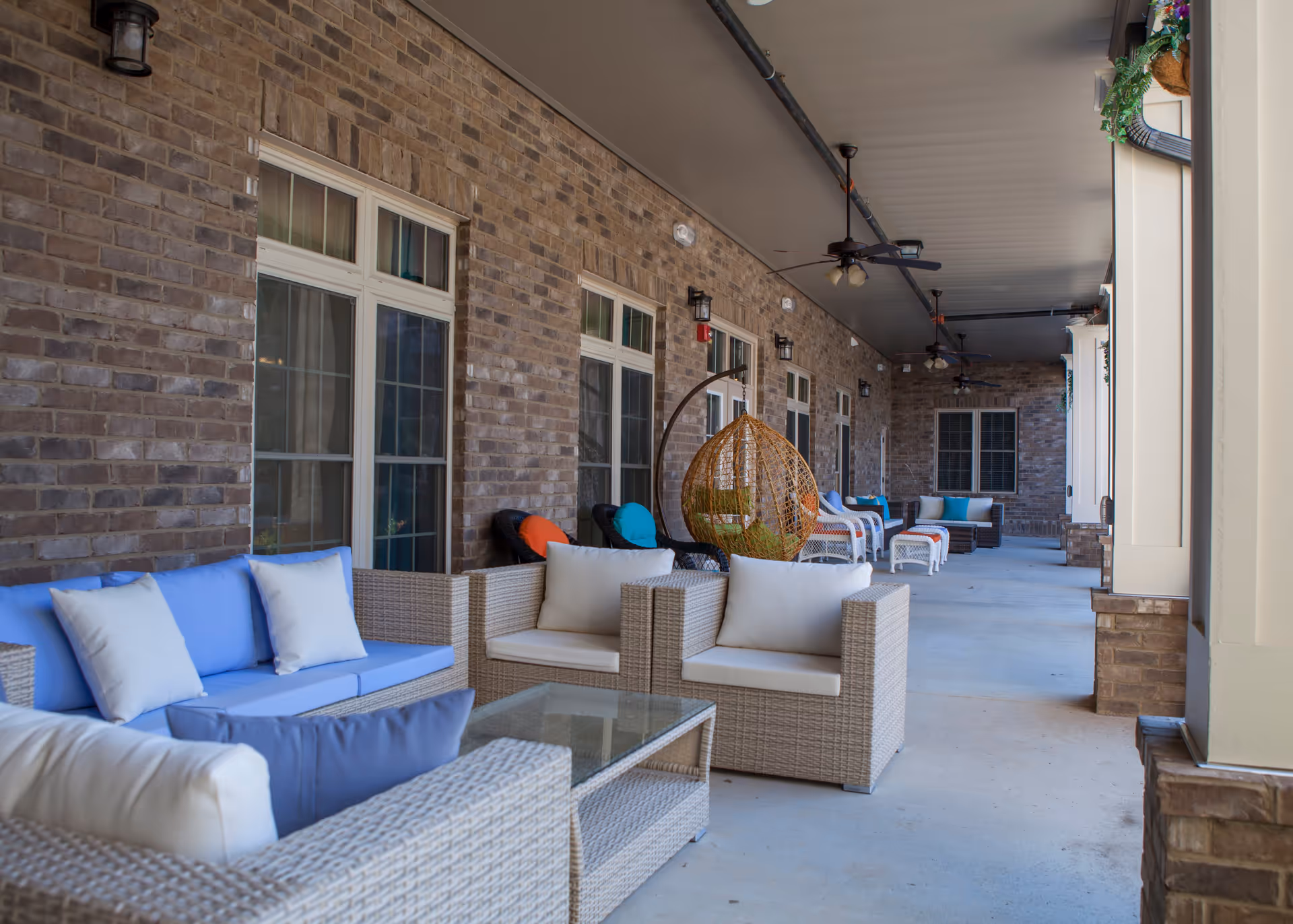 Covered outdoor patio area with multiple seating arrangements including wicker sofas and chairs with cushions in blue, white, and orange. The patio has a brick wall with windows and ceiling fans overhead. There is a hanging wicker egg chair and some greenery hanging from the ceiling.