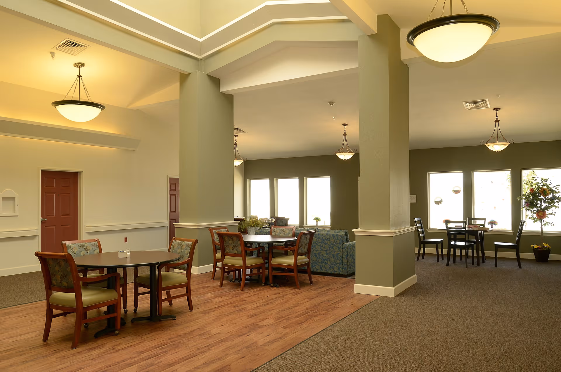 A spacious senior living common area with multiple round tables and chairs arranged for seating. The room features a mix of wood and carpet flooring, green walls with white trim, large windows allowing natural light, and ceiling light fixtures. There is a blue patterned sofa near the windows and a potted plant in the corner.