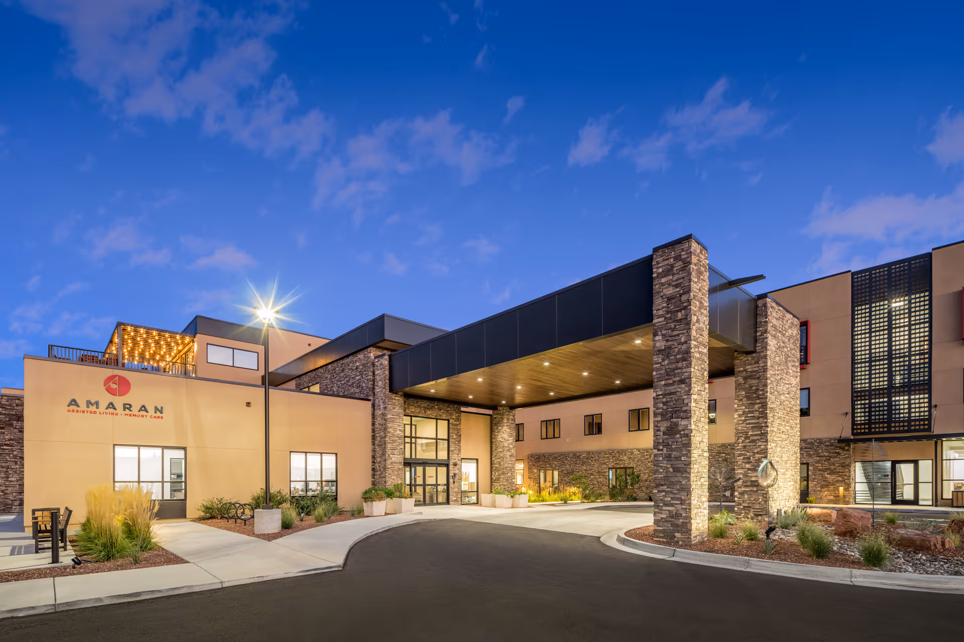 Exterior view of Amaran Senior Living facility at dusk, showing a modern building with stone pillars, large windows, and a covered entrance under a partly cloudy sky.