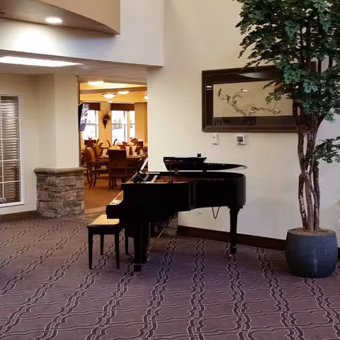 Interior view of a senior living facility featuring a black grand piano with a matching bench on a patterned carpet. To the right of the piano is a large potted plant. In the background, there is a dining area with tables and chairs, visible through an open doorway. A framed artwork hangs on the wall above the piano.