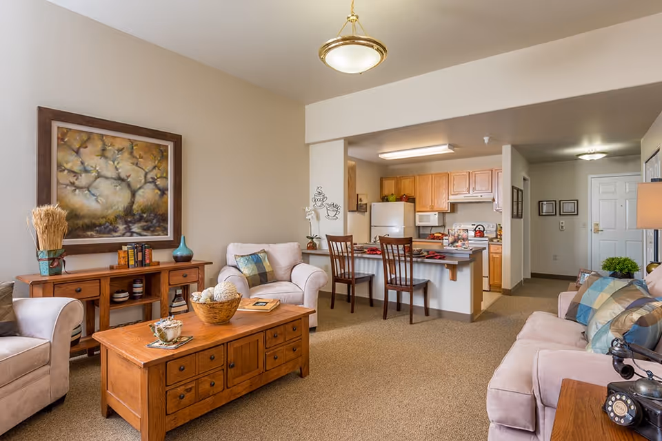 A cozy senior living apartment interior featuring a living room with beige armchairs and a sofa adorned with colorful pillows, a wooden coffee table and sideboard, a framed painting on the wall, and a kitchen area with wooden cabinets, a refrigerator, microwave, and a breakfast bar with two chairs.