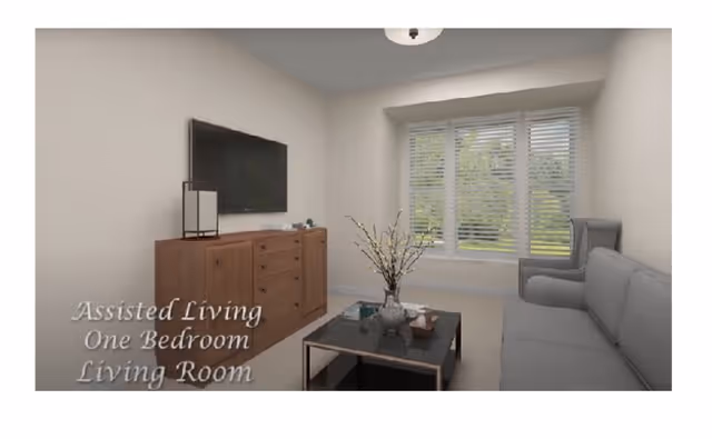 A living room in an assisted living one-bedroom unit featuring a gray sofa, a gray armchair, a wooden cabinet with a flat-screen TV mounted above it, a coffee table with decorative items, and a large window with white blinds showing greenery outside.