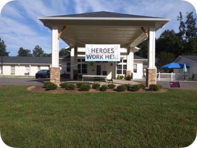 Front exterior view of Meadowview Nursing Center with a covered entrance supported by white columns with stone bases. A banner hanging from the entrance reads 'HEROES WORK HERE'. There is a green lawn in the foreground and trees in the background under a partly cloudy sky.
