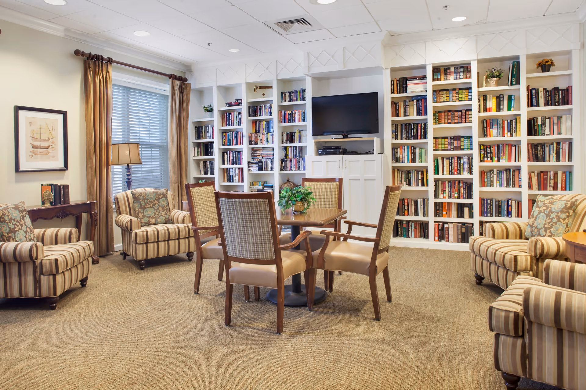 Comfortable community library/lounge with striped armchairs, a central table and chairs, and built-in bookshelves with a TV.