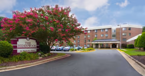 Exterior view of The Hidenwood Retirement Community building with a driveway leading to the entrance. A large tree with pink flowers is on the left side near a sign displaying the community's name and address. Several cars are parked near the building under a partly cloudy sky.
