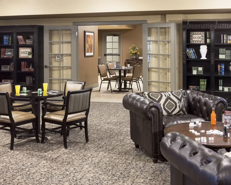 A cozy common area in a senior living facility featuring dark leather tufted sofas with patterned cushions, a round wooden table with dominoes and drinks, and a round dining table with four chairs. The room has carpeted flooring and is flanked by dark wooden bookshelves filled with books and decorative items. In the background, there is a hallway with additional seating and a door with glass panels.