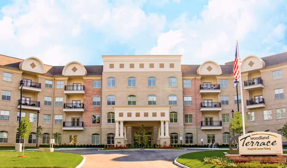 Front exterior view of Woodland Terrace of Carmel, a multi-story senior living facility with balconies, an entrance with columns, and a landscaped driveway with an American flag and a sign displaying the facility's name.