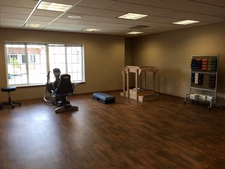 A spacious room with wooden flooring and beige walls, featuring exercise equipment including a recumbent bike, a wooden parallel bars structure, a step platform, and a rack holding various colored weights and exercise bands. A large window lets in natural light.