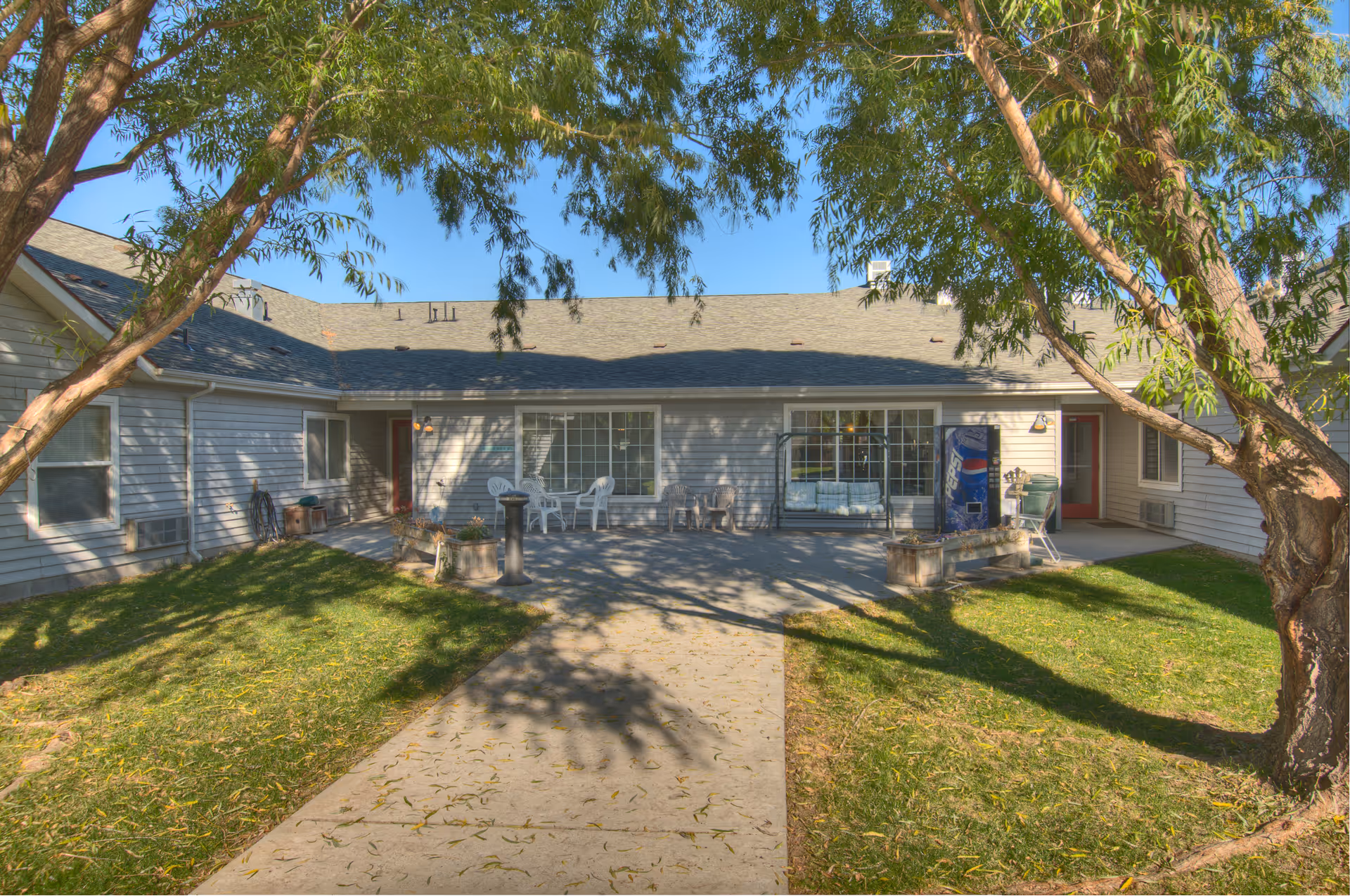 Courtyard and patio of a single-story assisted living building with chairs, a vending machine, and trees.