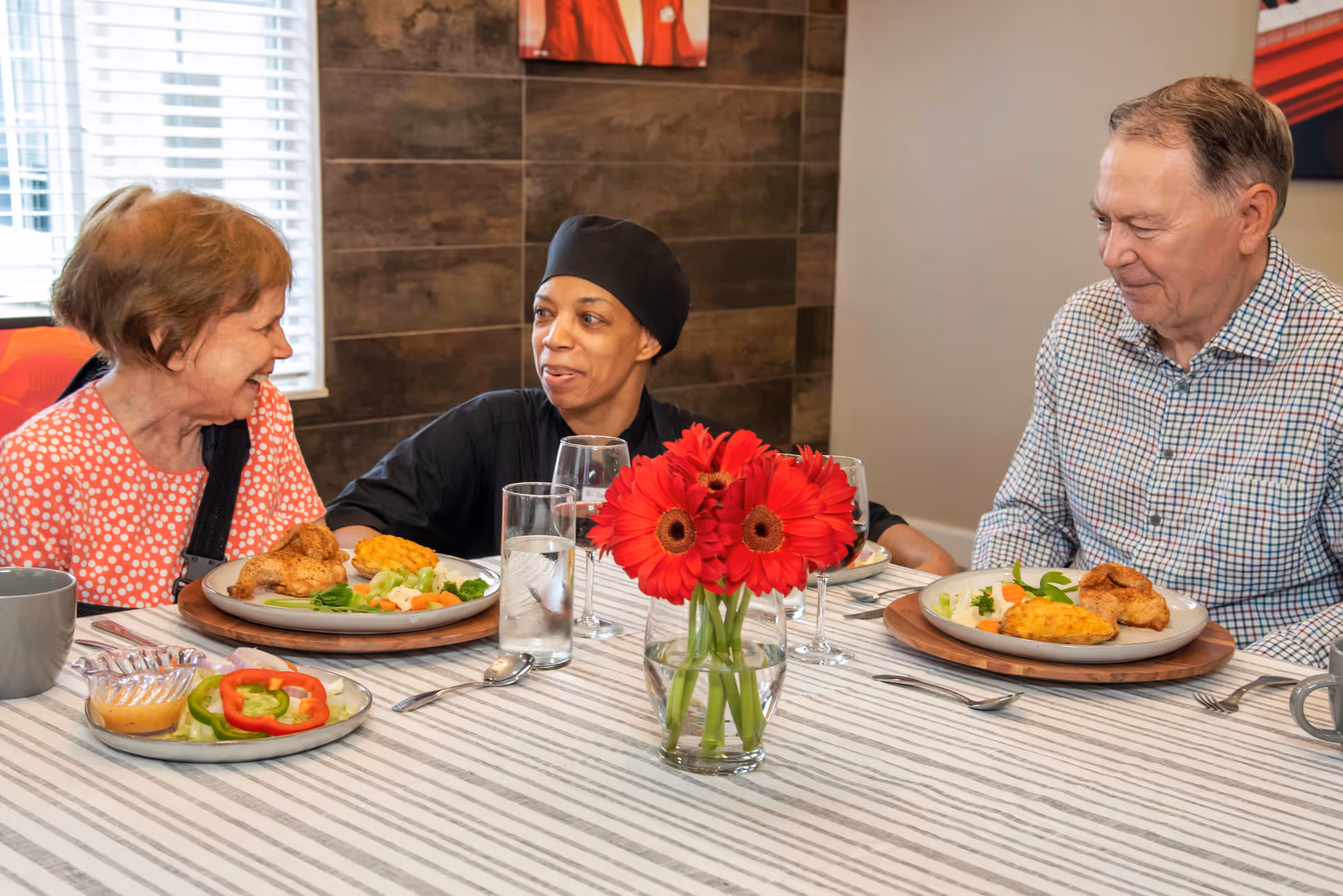 Two elderly individuals and a caregiver sitting at a dining table with plates of food, smiling and engaging in conversation. The table has a striped tablecloth, a vase with red flowers, and glasses of water.