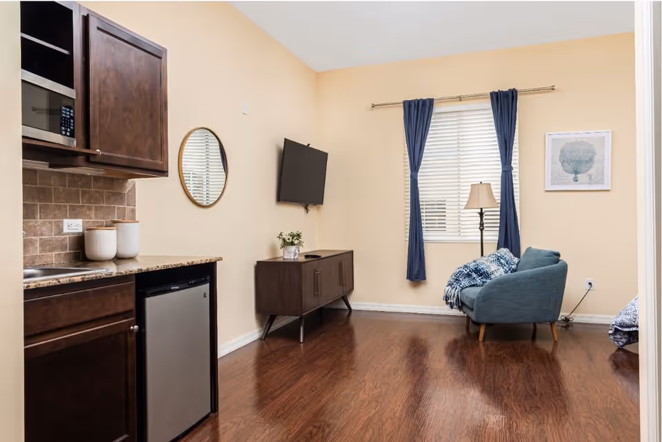 A cozy living area with wooden flooring featuring a small kitchenette with dark wood cabinets, a microwave, and a mini fridge on the left. A round mirror hangs on the wall above the counter. On the right side, there is a wall-mounted flat-screen TV above a wooden console table with a small plant. A window with blue curtains and white blinds is centered on the far wall, next to a floor lamp and a blue upholstered chair with a knitted throw blanket. A framed artwork hangs on the wall beside the window.