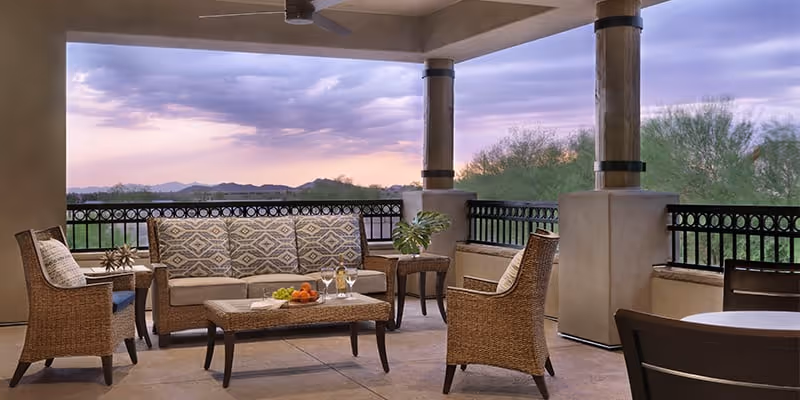 Covered outdoor patio area with wicker furniture including a sofa with patterned cushions, two armchairs, and a coffee table with fruit and drinks. The patio overlooks a scenic view of trees and distant mountains under a cloudy sky at sunset.