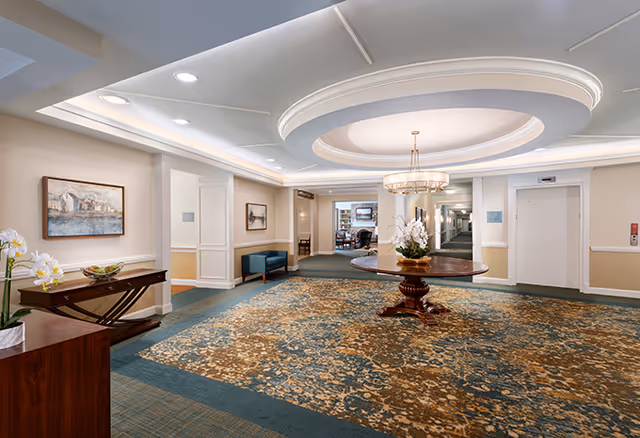 A spacious and well-lit senior living facility interior with a large patterned carpet, a round wooden table with a floral arrangement in the center, a chandelier hanging from a recessed ceiling, and seating areas along the walls. There are paintings on the walls and an elevator on the right side.