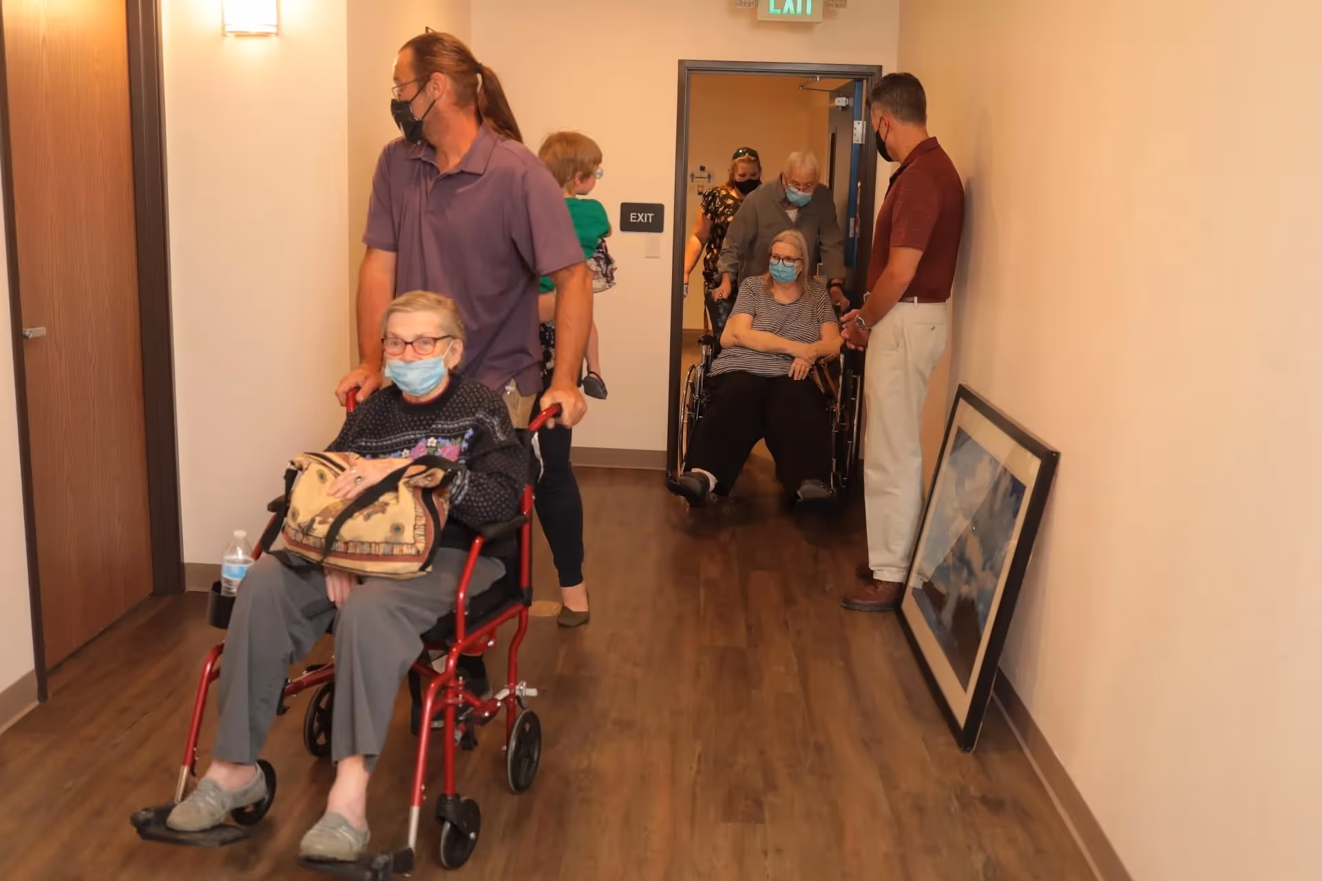 A hallway in an assisted living facility with two elderly women in wheelchairs being pushed by caregivers wearing face masks. Other people, including a child, are also present in the hallway. A framed picture is leaning against the wall on the right side.