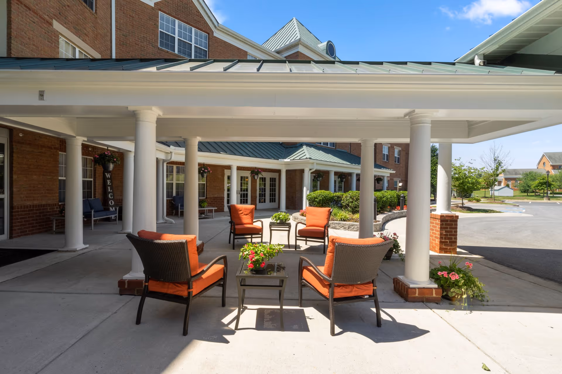 Outdoor covered seating area at a senior living facility with four cushioned chairs arranged around a small table with flowers. The building has brick walls, white columns, and a green metal roof. There are hanging flower baskets and a welcome sign near the entrance.