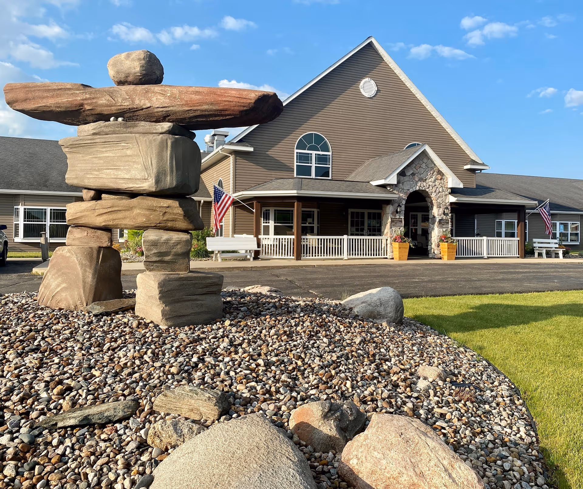 Front exterior view of StoneyBrook Suites Assisted Living building with a stone sculpture in the foreground, American flags on either side of the entrance, and a clear blue sky.