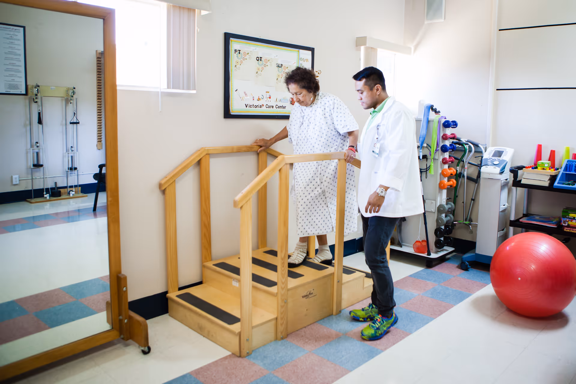 An elderly woman in a hospital gown is using a small wooden staircase with handrails for physical therapy, assisted by a healthcare professional in a white coat. The room contains exercise equipment, including weights and a large red exercise ball, with a mirror on one wall and a window letting in natural light.