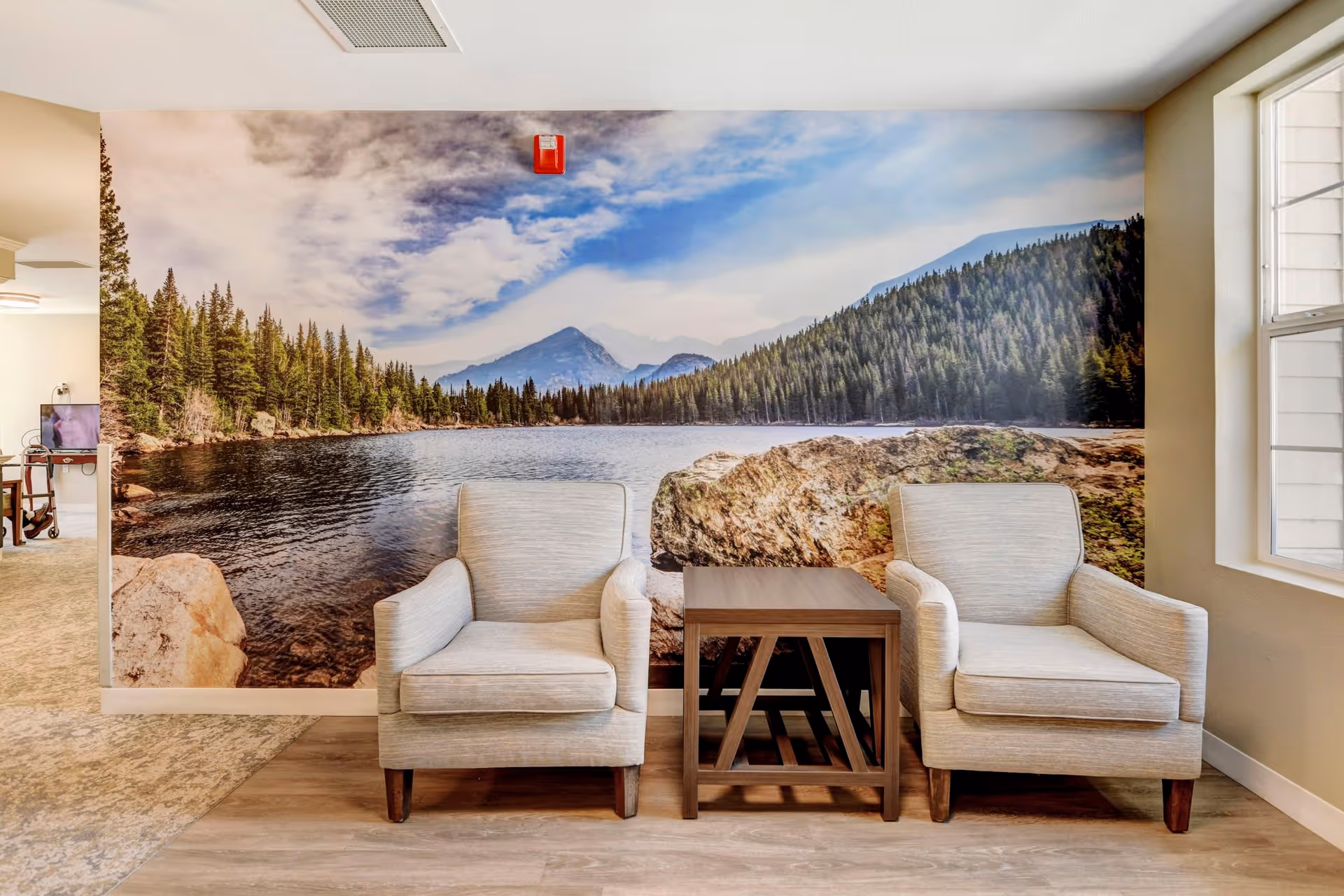 Two beige armchairs with wooden legs are placed on either side of a small wooden side table in a room. Behind the chairs is a large wall mural depicting a scenic lake surrounded by pine trees and mountains under a partly cloudy sky. To the right, there is a window letting in natural light, and to the left, part of another room with a TV and walker is visible.