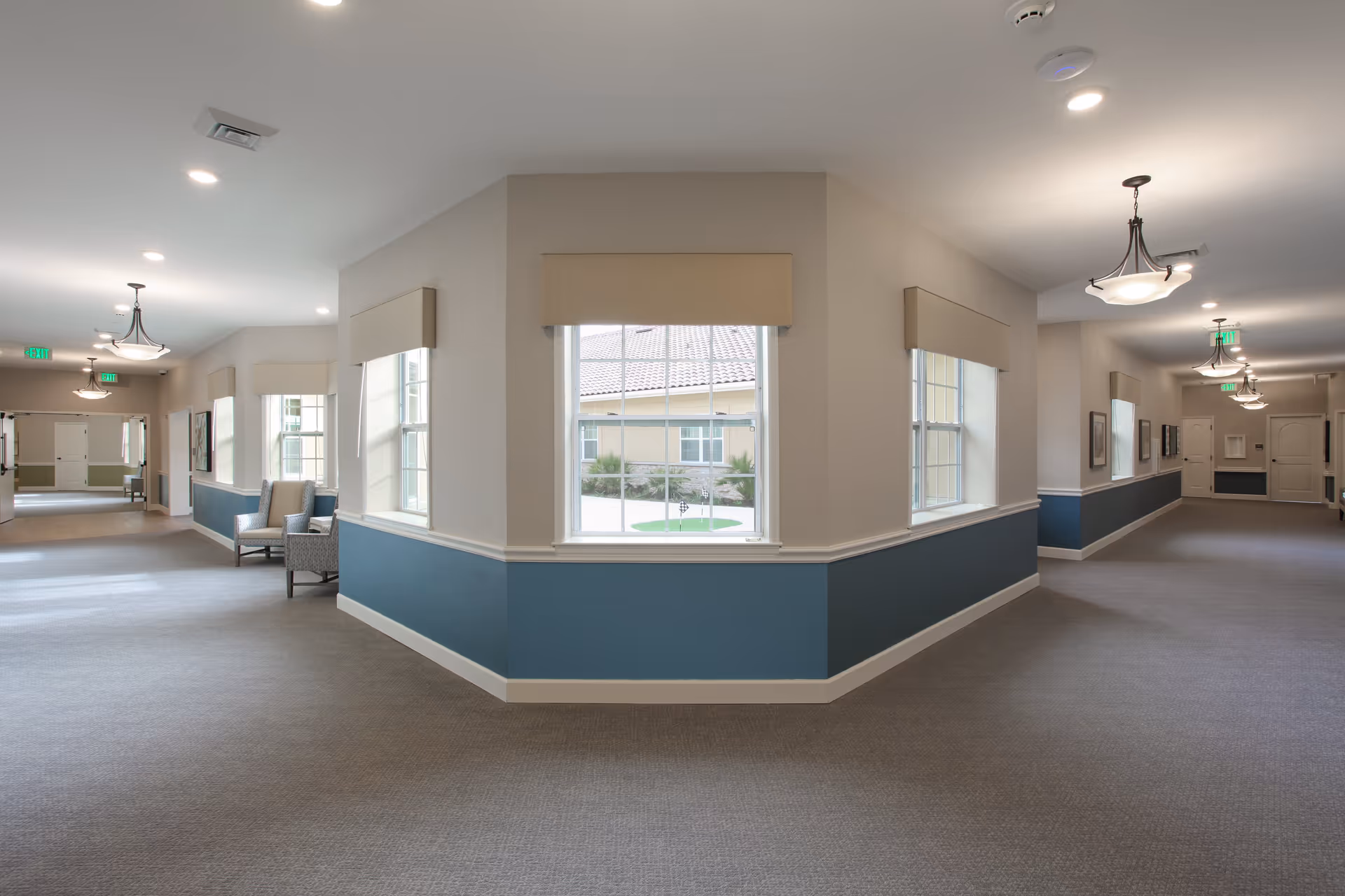 A spacious, well-lit hallway in a senior living facility with beige walls and blue wainscoting. The hallway features multiple windows with beige valances, ceiling lights, and a few chairs placed along the walls. The carpet is gray, and there are exit signs visible at both ends of the corridor.