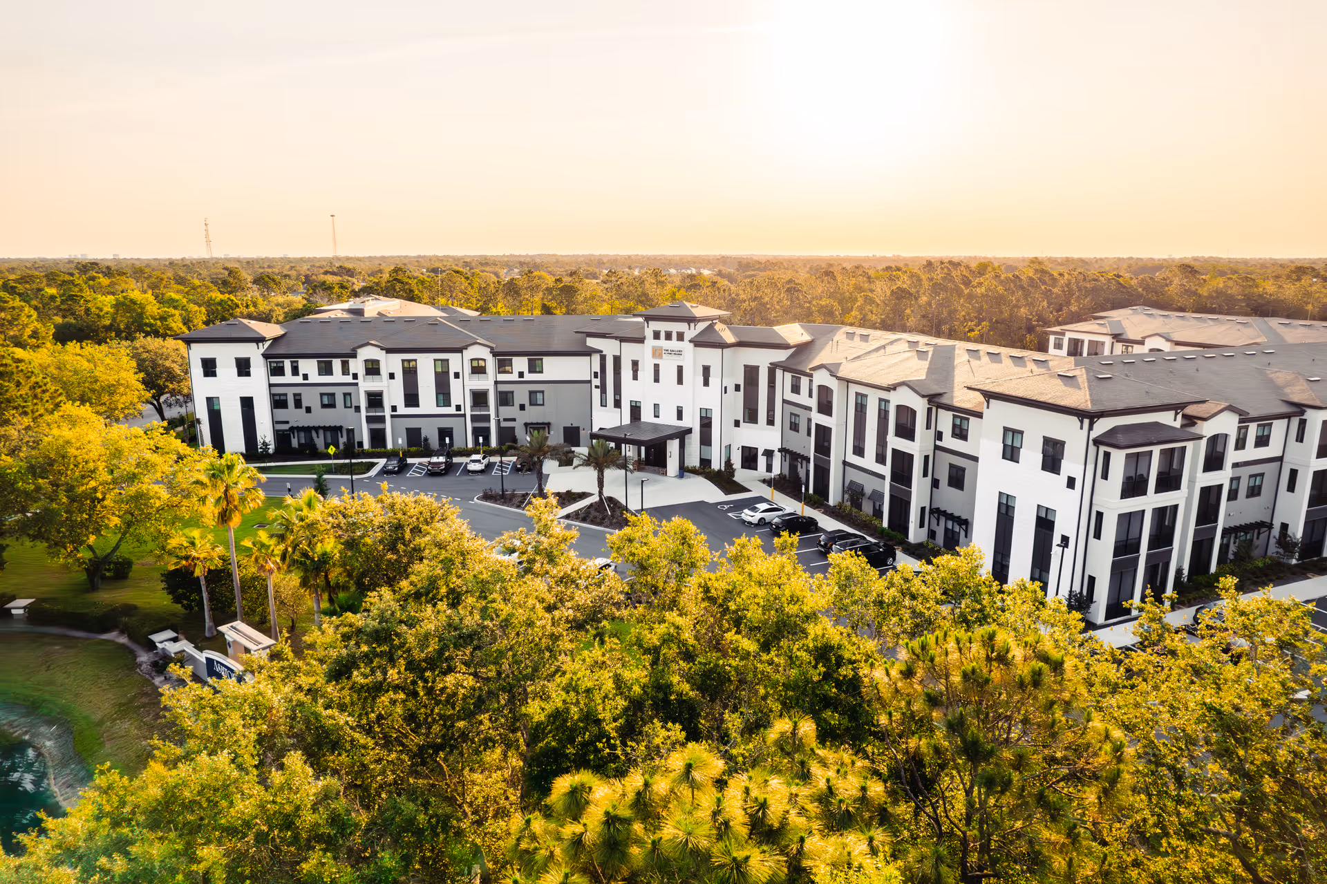 Aerial view of a large three-story senior living building surrounded by trees, parking areas, and a pond at sunset.