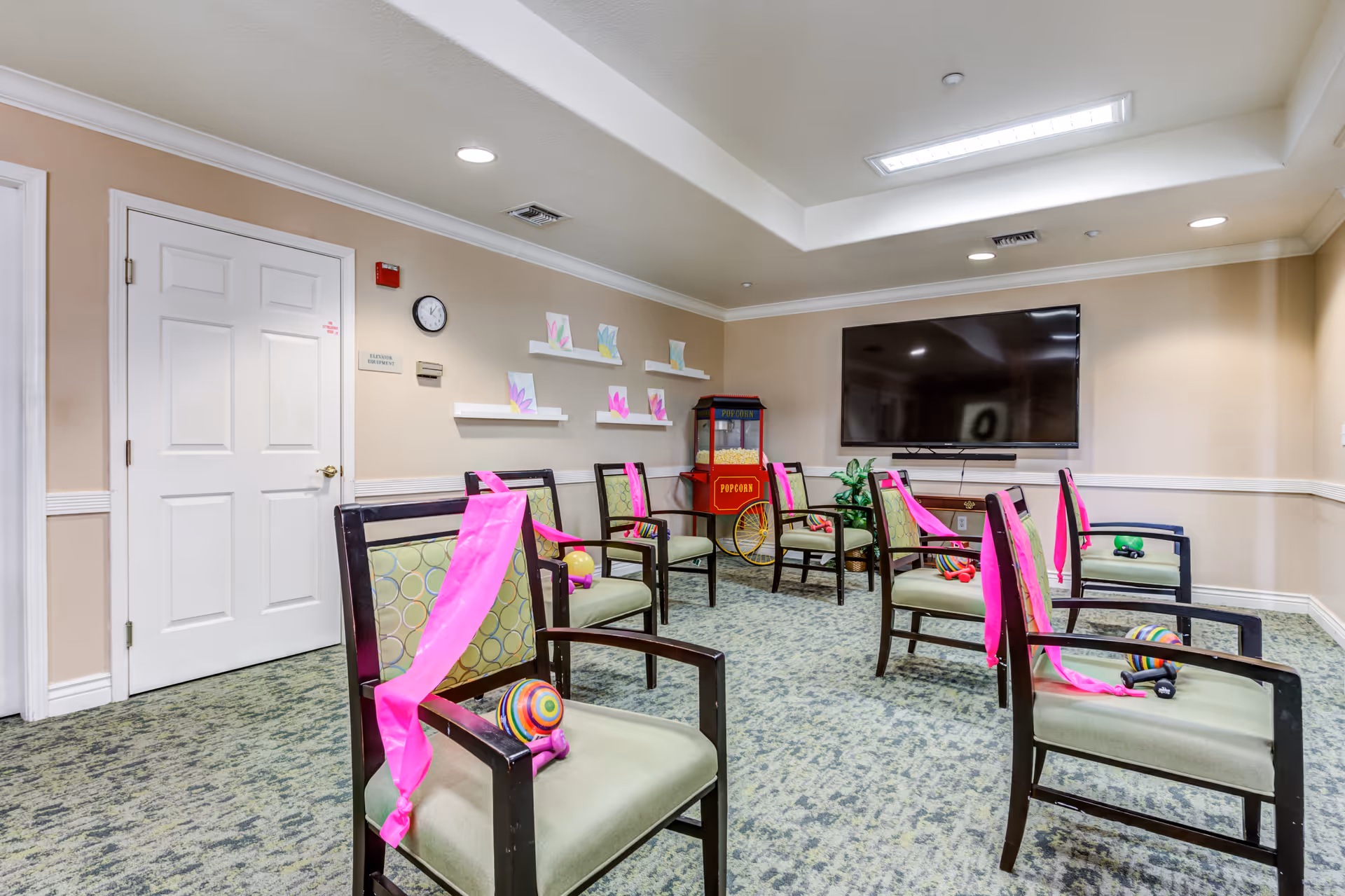 A room with several chairs arranged in a circle, each chair has a pink ribbon tied to the back and a colorful ball placed on the seat. The room has beige walls, a carpeted floor, a large flat-screen TV mounted on the wall, a popcorn machine in the corner, and some children's artwork displayed on white shelves.