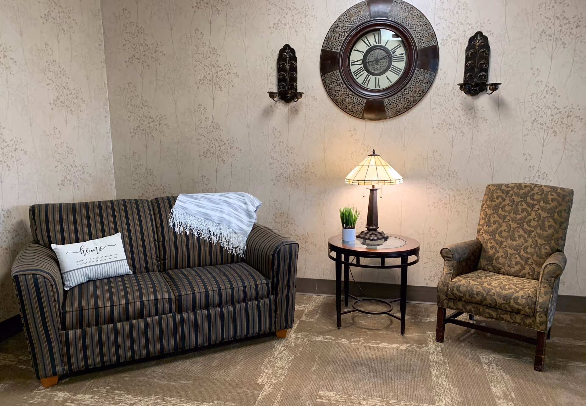 A cozy sitting area with a striped couch featuring a decorative pillow and a throw blanket, a patterned armchair, and a round side table with a lamp and a small potted plant. Above the table is a large decorative wall clock flanked by two wall sconces. The walls have a subtle floral pattern wallpaper.
