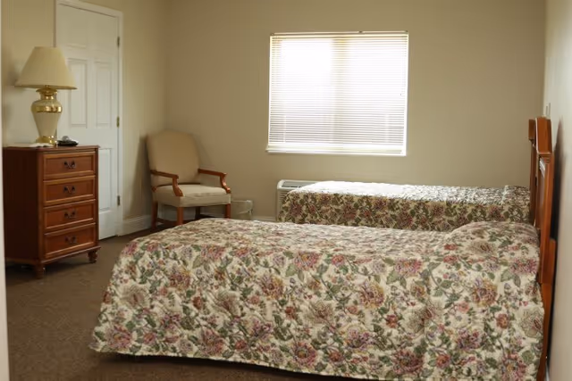 A simple bedroom with two twin beds covered in floral bedspreads, a wooden dresser with a lamp on top, a beige armchair, and a window with closed blinds letting in natural light.