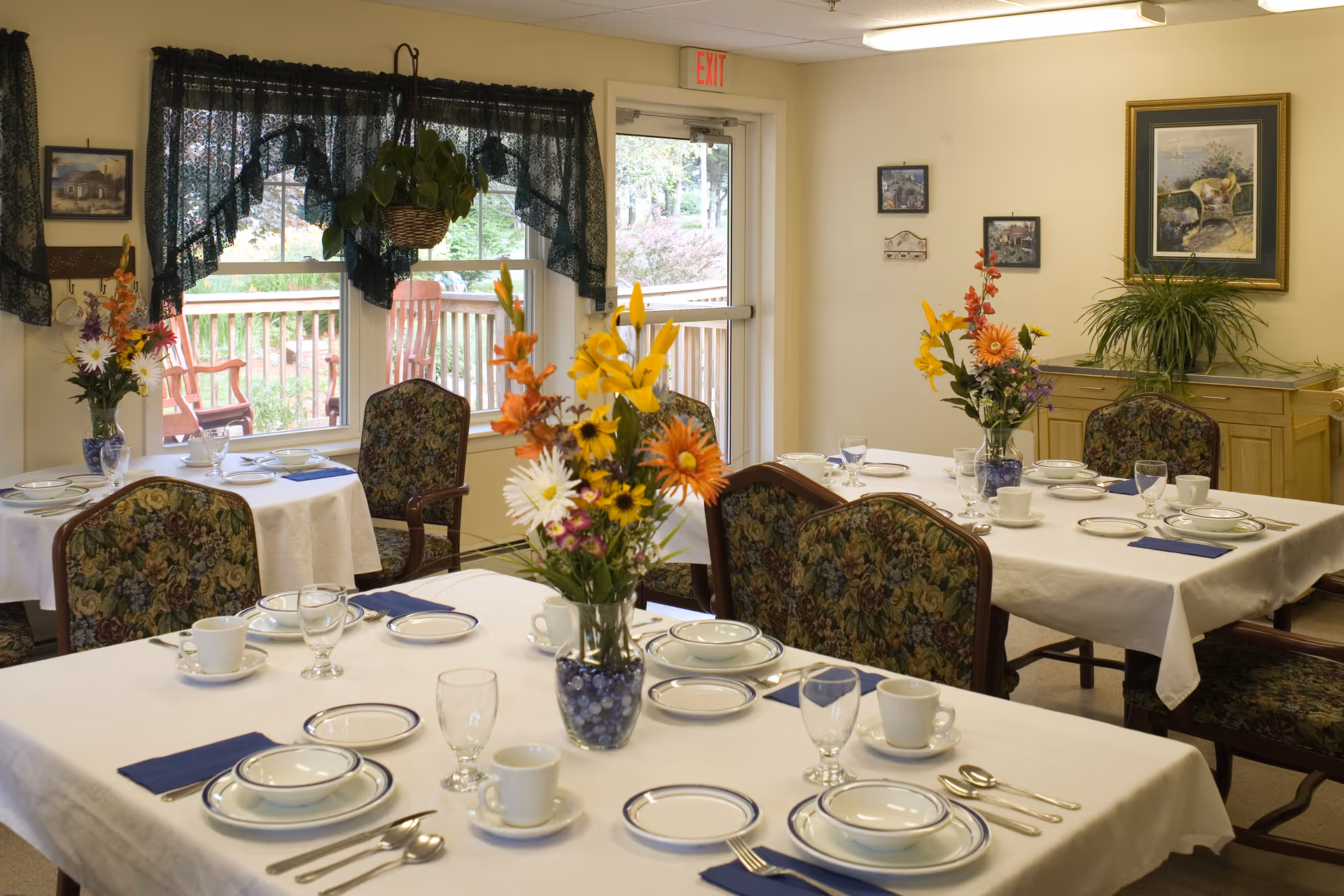 Sunlit dining room with tables set for a meal, floral centerpieces, and patterned chairs near windows and an exit door.