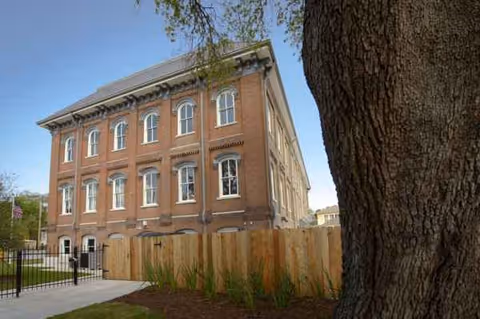 Brick multi-story building with arched windows, a wooden fence, and a large tree in the foreground.