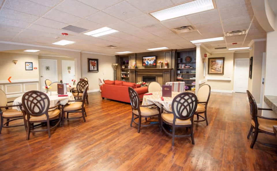 Open communal dining and lounge area with round dining tables and chairs, wood floors, red sofas, and a fireplace with a TV.