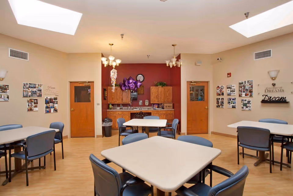 A well-lit dining room with several square tables surrounded by blue chairs. The walls are decorated with photo collages and inspirational words like 'Dream' and 'Gallery of Dreams'. There are two wooden doors and a small kitchenette area with wooden cabinets and a clock on a red accent wall. The ceiling has two skylights and two chandeliers.