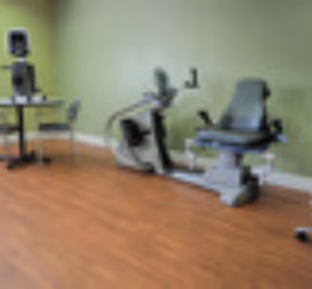 A small exercise/therapy room with a recumbent bike and fitness equipment against a green wall and wood floor.