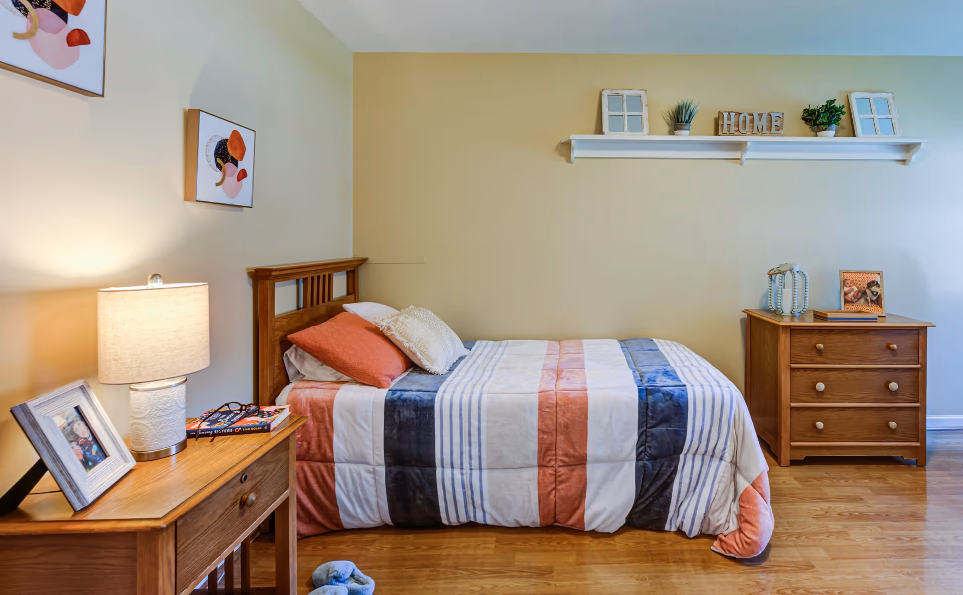 A cozy bedroom with a single wooden bed featuring a striped comforter in shades of white, blue, and coral. The bed has two pillows, one coral and one white. To the left of the bed is a wooden nightstand with a lamp, a framed photo, and some books. On the right side of the room is a wooden dresser with decorative items and a framed photo on top. Above the bed, a white wall shelf holds small plants, decorative window frames, and wooden letters spelling 'HOME'. The room has light-colored walls and wooden flooring.