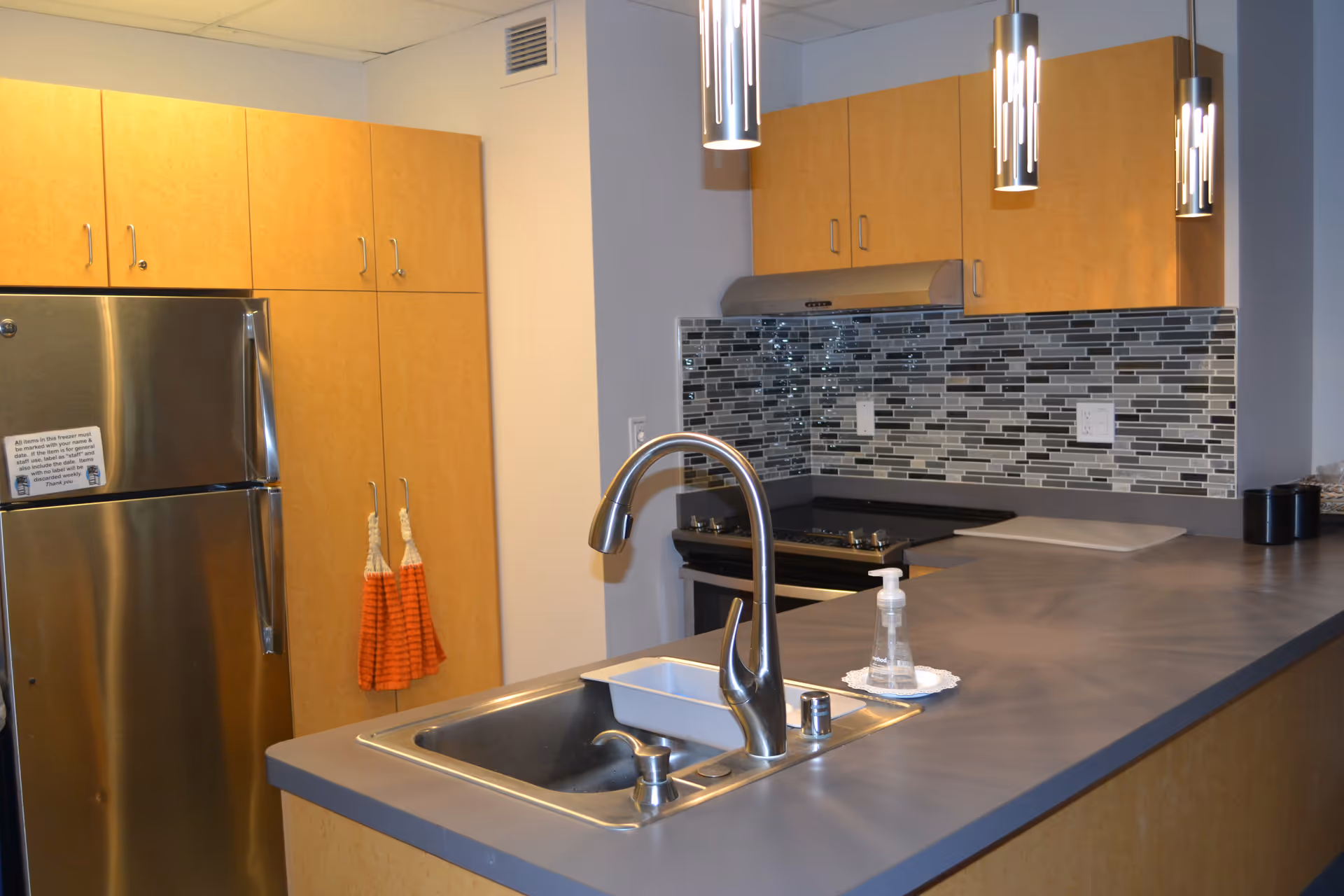 Modern kitchen area with stainless steel refrigerator, light wood cabinets, a gray countertop with a built-in sink and faucet, a stove with a stainless steel hood, and three hanging pendant lights. There is a bottle of hand soap on a small doily next to the sink and two orange hand towels hanging on cabinet handles.
