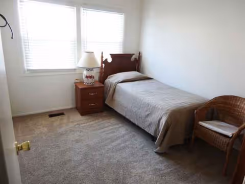Small bedroom with single bed, nightstand, lamp, and wicker chair by window.