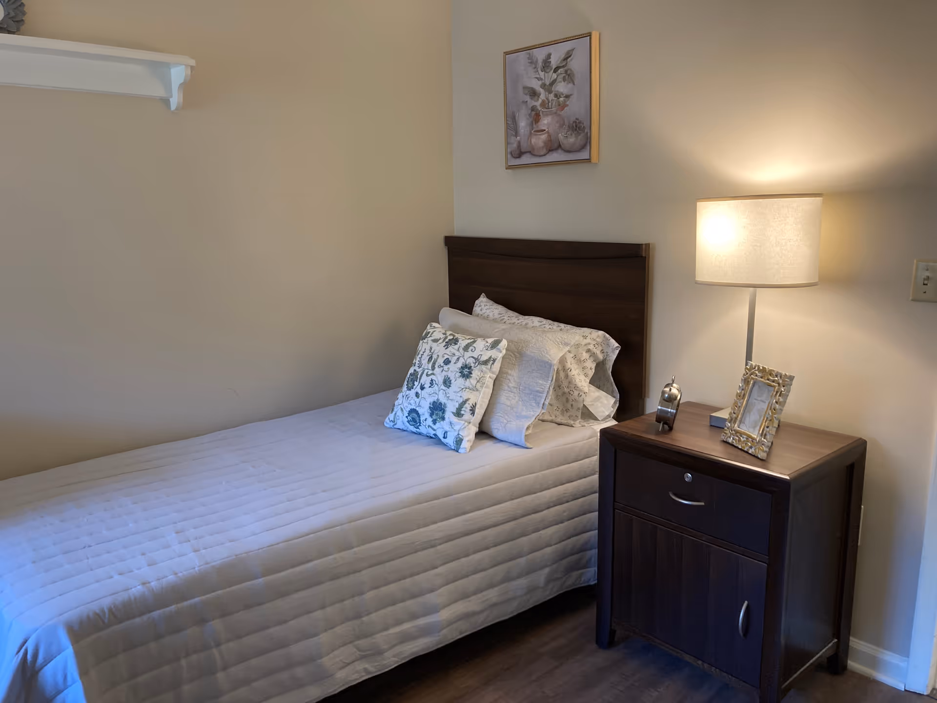 A small bedroom with a single bed covered in a white quilt and floral pillows. Next to the bed is a dark wooden nightstand with a lamp, a small clock, and a decorative picture frame. A framed floral painting hangs on the wall above the bed.
