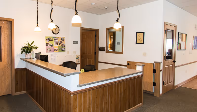 Wood-paneled reception desk with pendant lights, chairs, and doors in a senior living facility interior.