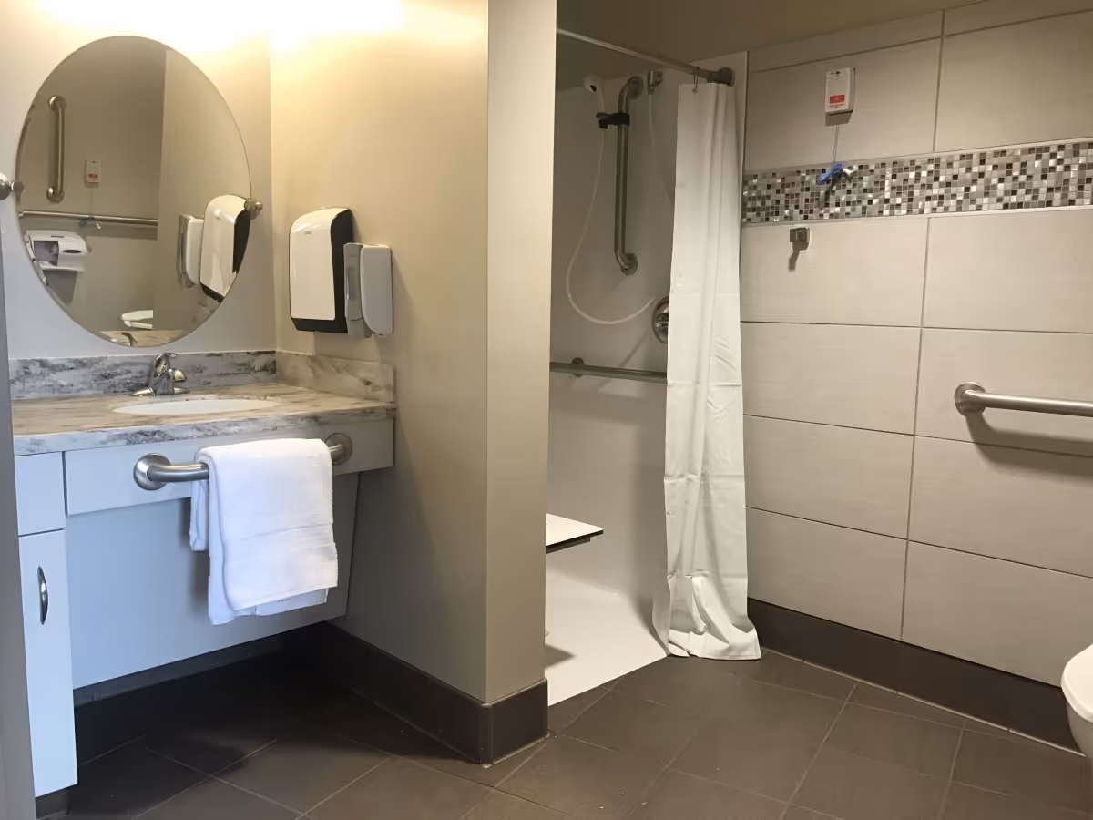 A clean, accessible bathroom with a sink and countertop on the left, a round mirror above the sink, a towel hanging on a grab bar, and a walk-in shower with a white curtain and grab bars on the right. The walls are tiled with a decorative mosaic strip, and the floor is tiled in a dark color.