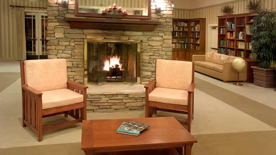 A cozy living room area with two wooden armchairs with beige cushions facing a wooden coffee table that has a book on it. Behind the chairs is a stone fireplace with a fire burning inside. To the right, there is a beige sofa, bookshelves filled with books, a globe on a stand, and a large potted plant. The walls have striped wallpaper and there is a mirror above the fireplace.