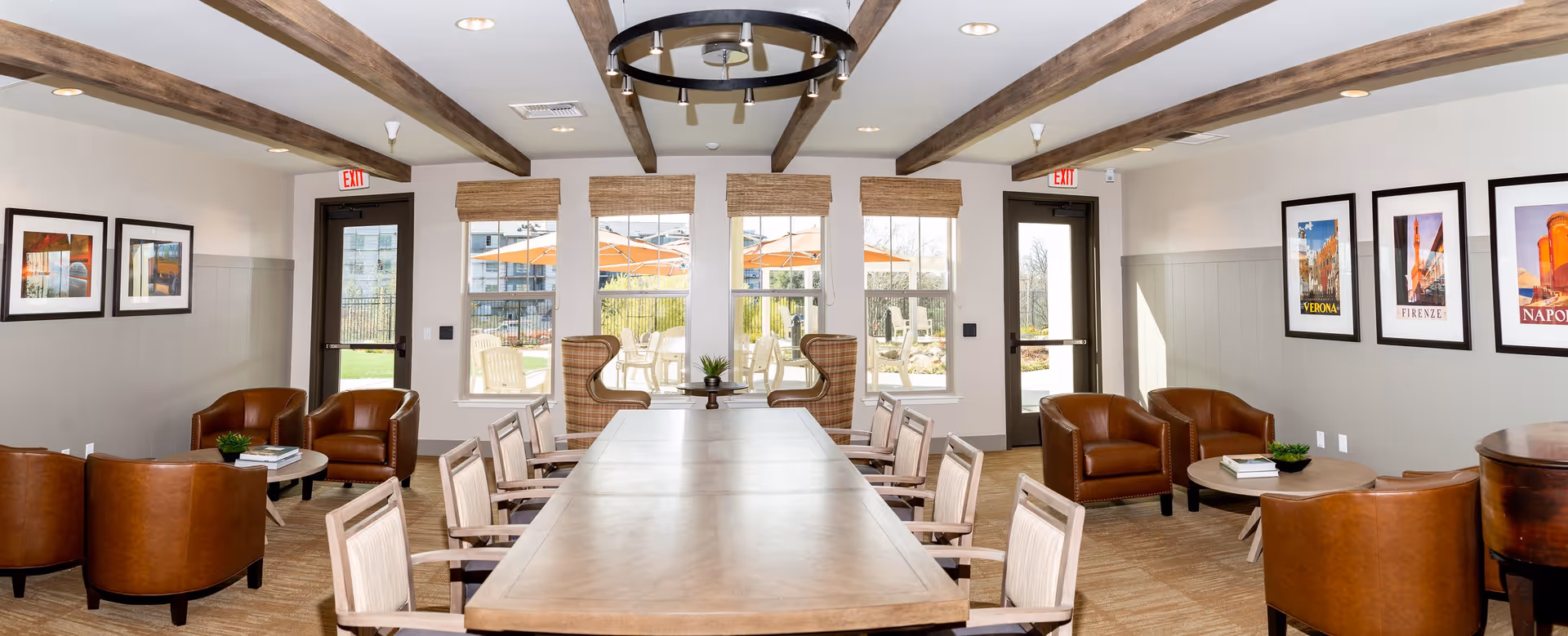 A spacious, well-lit common area with a long wooden table surrounded by chairs in the center. The room features exposed wooden ceiling beams, leather armchairs arranged around small round tables, and large windows with woven shades that look out onto an outdoor patio with umbrellas and chairs. The walls are decorated with framed pictures of various cityscapes.