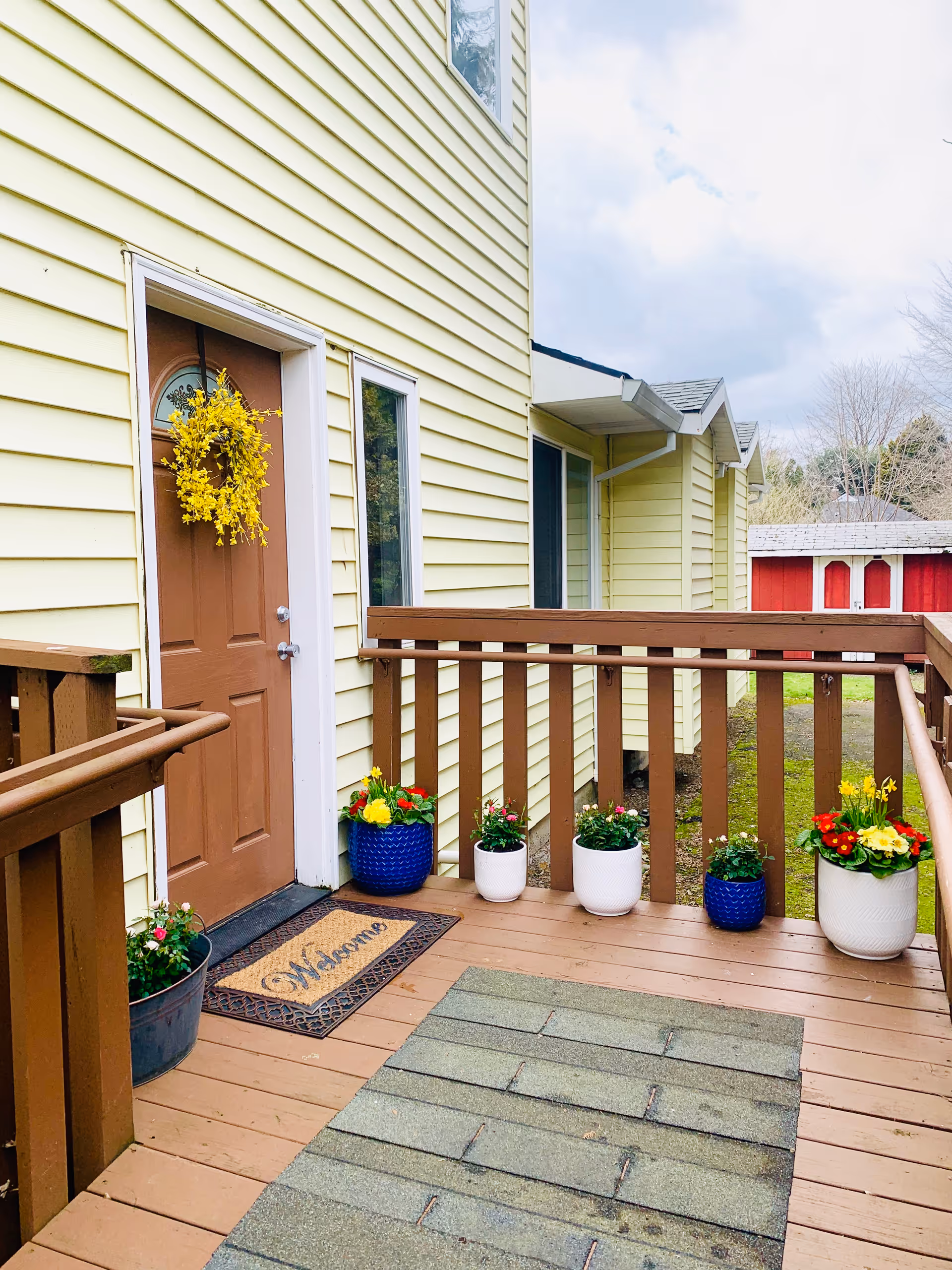 A wooden porch with a brown door decorated with a yellow flower wreath. The porch has a welcome mat and several flower pots with colorful flowers placed along the railing. The building exterior is light yellow with white trim, and there is a red shed visible in the background.