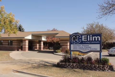 Exterior view of Fargo Elim Senior Living Community building with a clear blue sky. The building has a brick facade with a covered entrance and a driveway leading up to it. In front of the building is a sign that reads 'Elim A Caring Community' with additional text listing Pioneer House, Elim Care Center, and Elim Children's Center. There are trees with autumn foliage and a parked car visible on the right side.