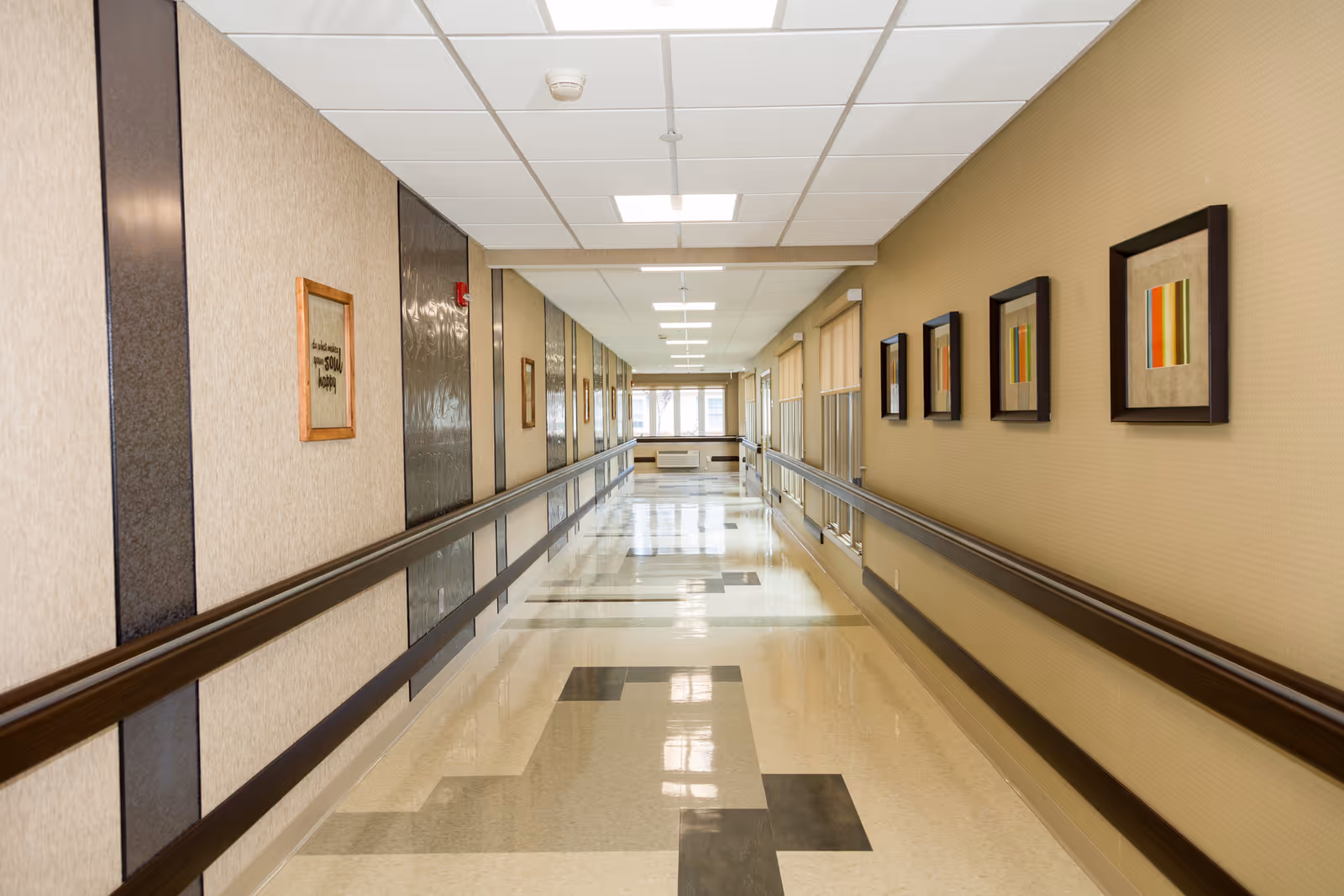 Long, well-lit interior hallway with handrails on both sides, framed artwork, and a patterned tile floor.