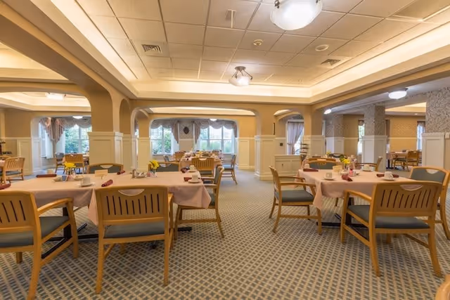 A spacious dining room with multiple wooden tables and chairs arranged neatly. Each table is covered with a light pink tablecloth and set with cups, saucers, and napkins. The room features large windows with curtains, soft lighting from ceiling fixtures, and a patterned carpet floor.