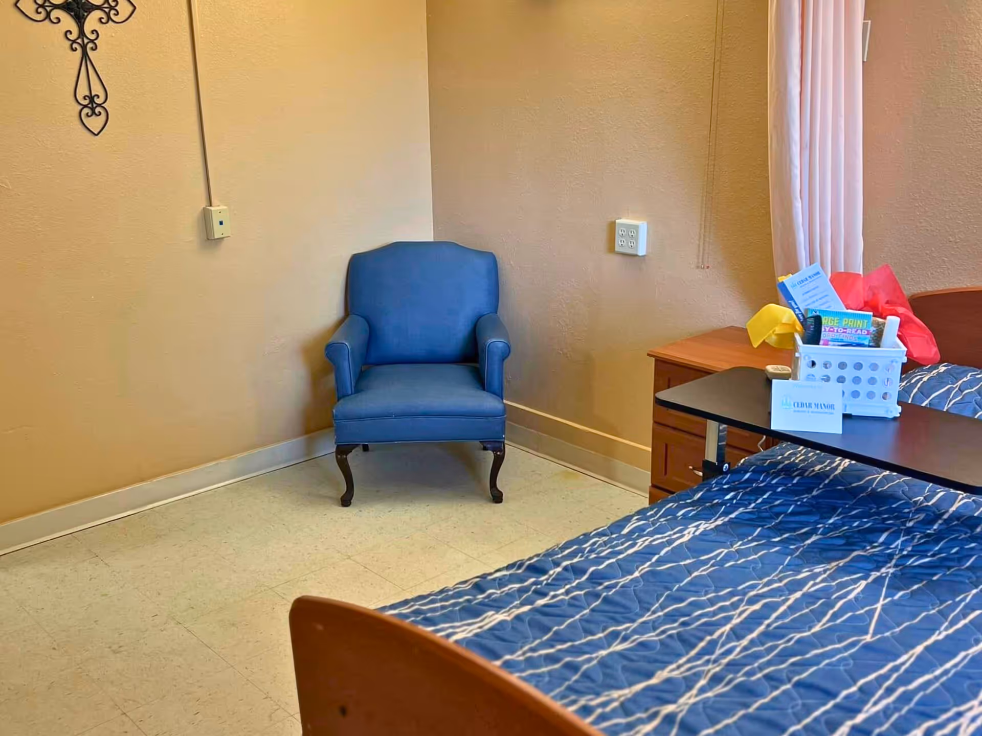 A nursing facility bedroom corner with a blue upholstered armchair next to a bed and a bedside table holding a basket and papers.