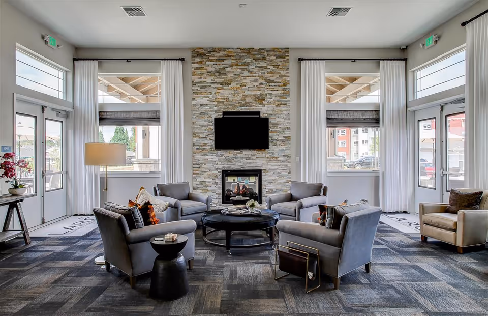 A modern living room with four gray armchairs arranged around a round black coffee table. Behind the seating area is a stone accent wall with a mounted flat-screen TV above a fireplace. Large windows with white curtains flank the fireplace, allowing natural light to fill the room. There are glass doors on both sides leading outside, and a floor lamp and side tables with decorative items complete the cozy setting.