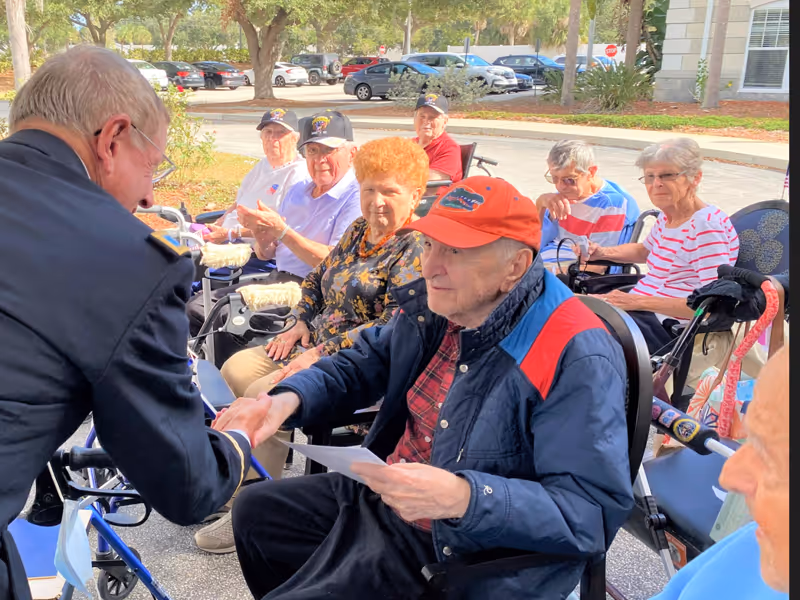 A group of elderly people sitting outdoors in wheelchairs and chairs, with one elderly man in a red cap shaking hands with a man in a dark uniform. Trees, parked cars, and a building are visible in the background.