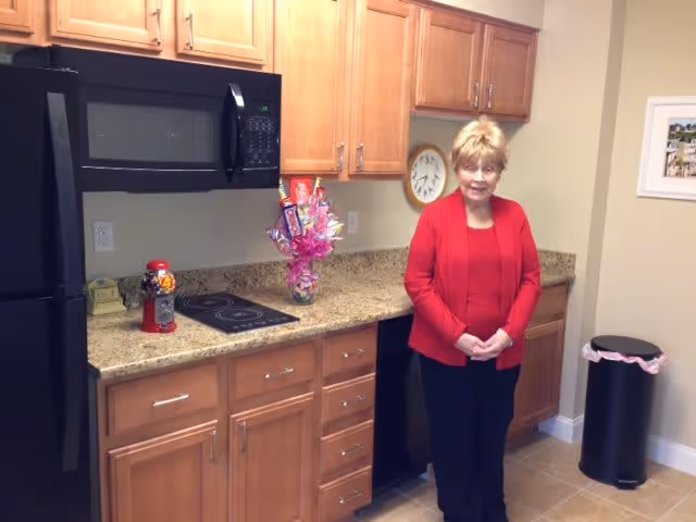An elderly woman wearing a red jacket and black pants stands in a kitchen with wooden cabinets, a granite countertop, a black microwave, and a black refrigerator. On the countertop, there is a small red gumball machine and a colorful candy bouquet. A wall clock and a framed picture are visible on the wall, and a black trash can with a pink liner is in the corner.