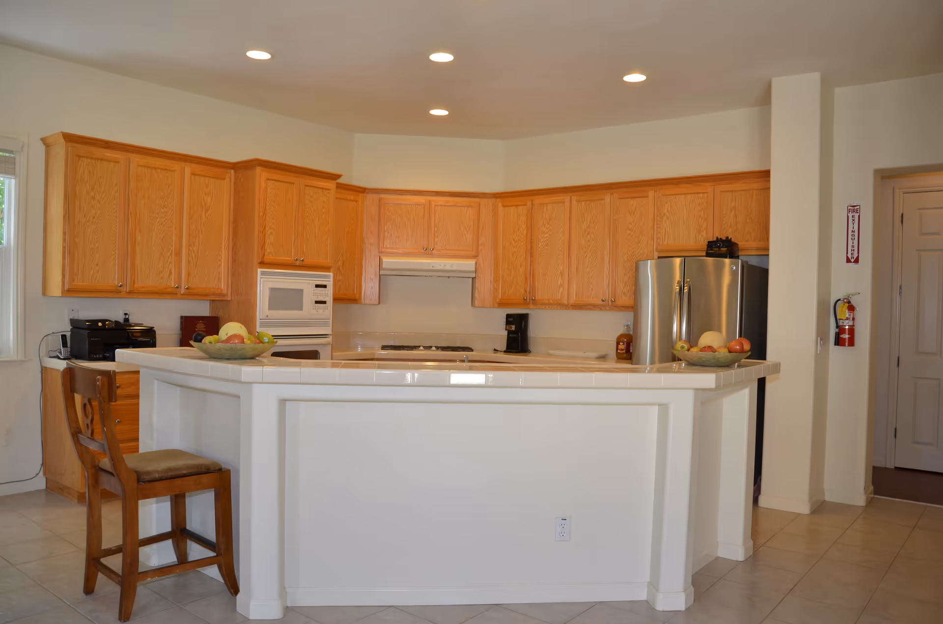A clean and spacious kitchen with wooden cabinets, a white tiled countertop island with a wooden chair, a stainless steel refrigerator, a built-in microwave, a coffee maker, and bowls of fruit on the counter. The kitchen has recessed ceiling lights and a fire extinguisher mounted on the wall near a doorway.